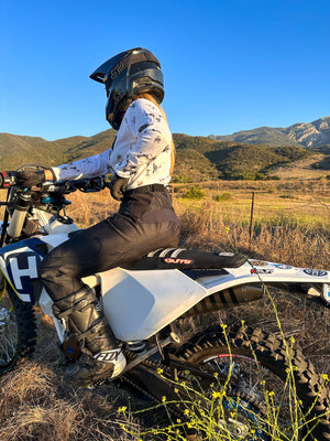 Woman sitting on her dirt bike wearing womens black dirt bike pants and a white bull skull jersey