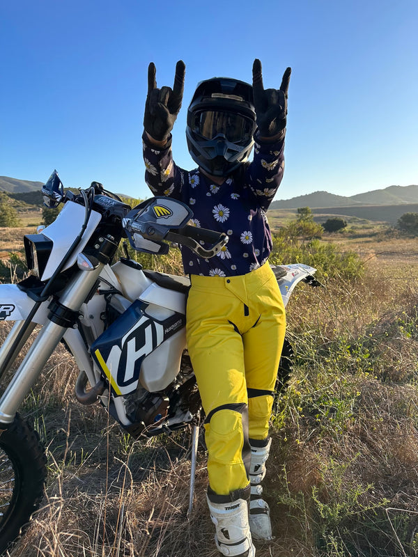 Woman standing next to her dirt bike wearing a navy blue and daisy dirt bike jersey and bright yellow womens dirt bike pants