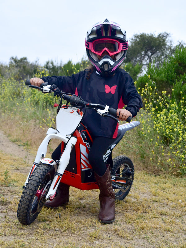 A girl sitting on a dirt bike wearing a grey and black camo jersey with hot pink accents.
