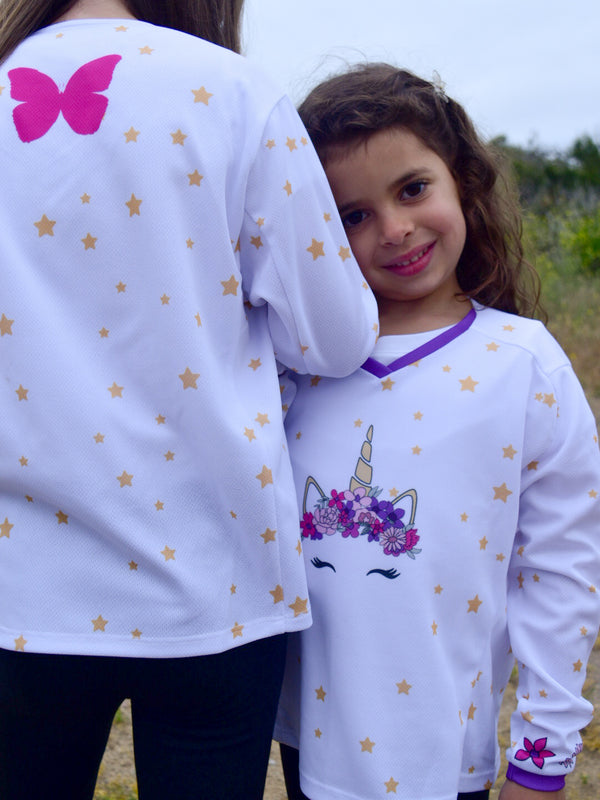 Two sisters wearing a white dirt bike jersey with a unicorn wearing a flower crown on the front, surrounded by stars
