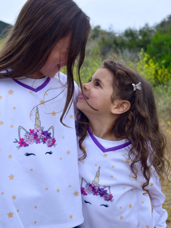 Two sisters wearing a white dirt bike jersey with a unicorn wearing a flower crown on the front, surrounded by stars