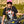 Load image into Gallery viewer, Woman wearing a black shirt with a flower skull on the front. She&#39;s wearing a neon pink trucker hat, standing behind her dirt bike.

