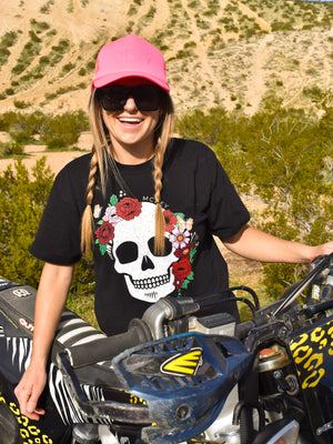 Woman wearing a black shirt with a flower skull on the front. She's wearing a neon pink trucker hat, standing behind her dirt bike.