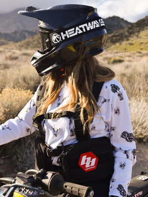 Woman wearing a white jersey with grey bull skulls on it, Heat Wave goggles, and a black pack. She is sitting on her dirt bike in the mountains.
