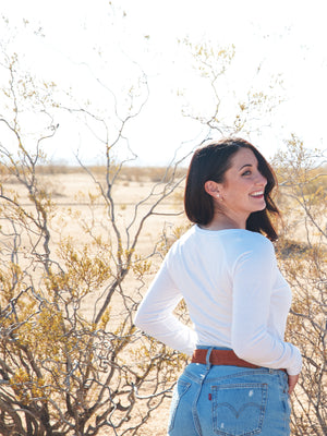 Woman wearing a white long sleeve shirt with small butterfly on the chest. She is smiling while standing in the desert. 