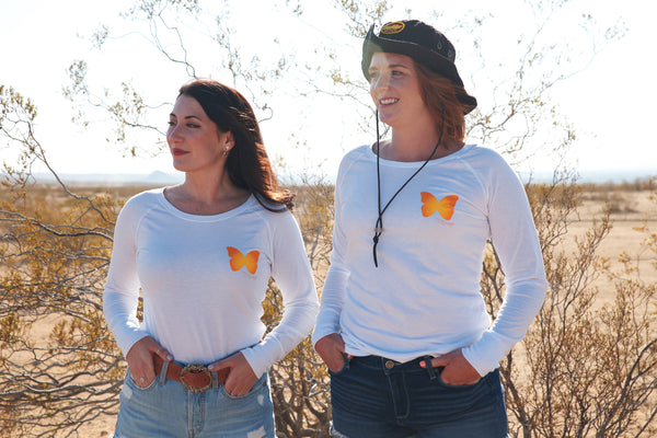 Two women wearing a white long sleeve shirt with small butterfly on the chest. They are smiling while standing in the desert. 