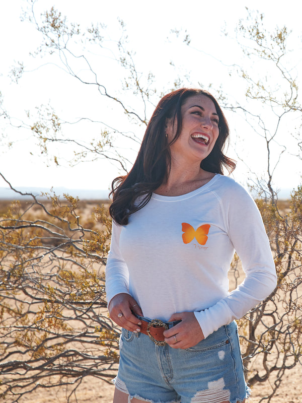Woman wearing a white long sleeve shirt with small butterfly on the chest. She is laughing while standing in the desert. 