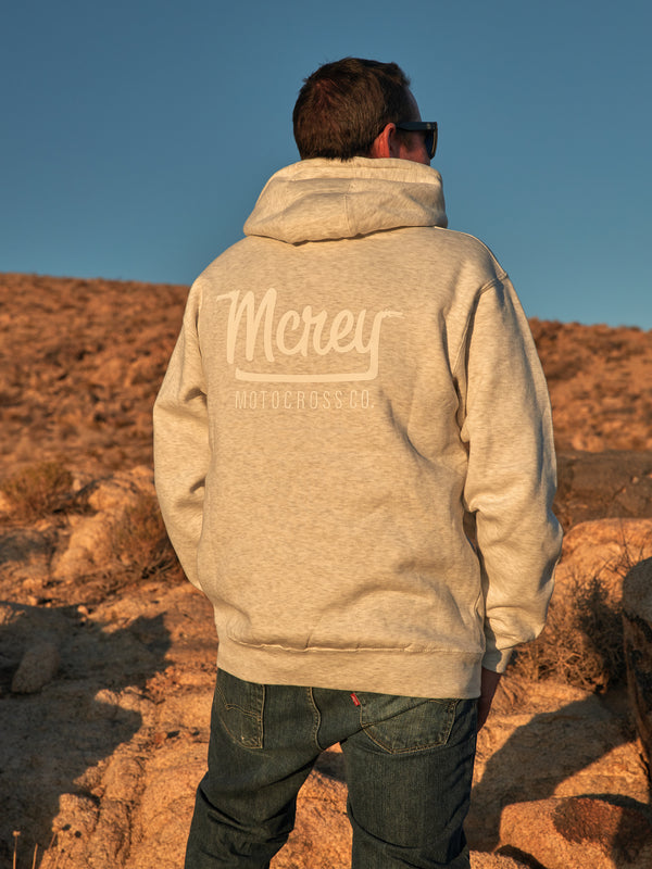 A man standing on a rock in the desert. He is wearing a heather oatmeal-colored hoodie sweatshirt with white logo on the back. Unisex dirt bike apparel.