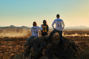 Three friends sit on a rock in the desert, watching an off-road vehicle drive by. They are wearing MCREY long sleeve shirts.