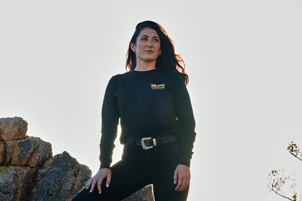 A woman stands on a rock in the desert. The sun is setting behind her. She is wearing a black long sleeve shirt with MCREY MOTOCROSS CO on the front.