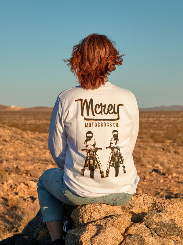 A woman is sitting on a rock in the desert. She is wearing a white long sleeve shirt with MCREY MOTOCROSS CO on the back and two girls sitting on dirt bikes.