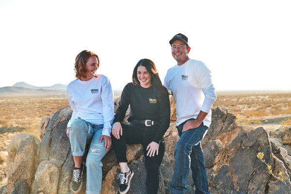 Three friends sit on a rock in the desert. They are laughing. They are wearing motocross long sleeve shirts.