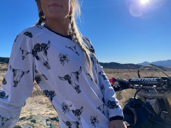 Woman wearing MCREY Bullish jersey sitting on a dirt bike. White with grey bull skulls and flowers all over.