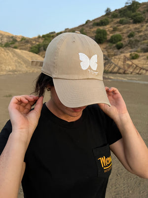 Woman with brown hair wearing a khaki-colored baseball-style cap with a white butterfly stitched on the front