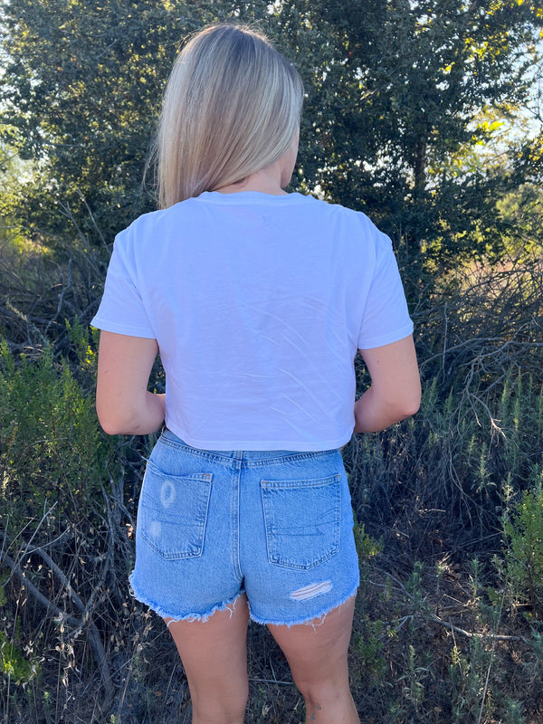 Blonde woman wearing a white crop shirt with black writing that says designed to get dirty standing in front of a tree