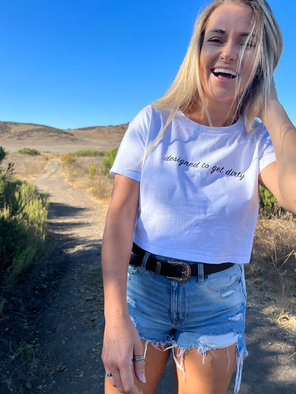 Blonde woman wearing a white crop shirt with black writing that says designed to get dirty in the middle of a field