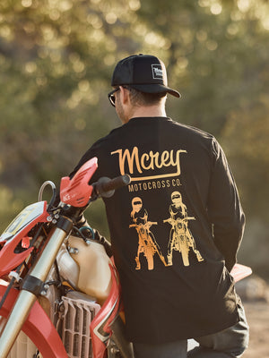 A man standing next to his dirt bike wearing a unisex black long sleeve shirt with two girls riding motorcycles