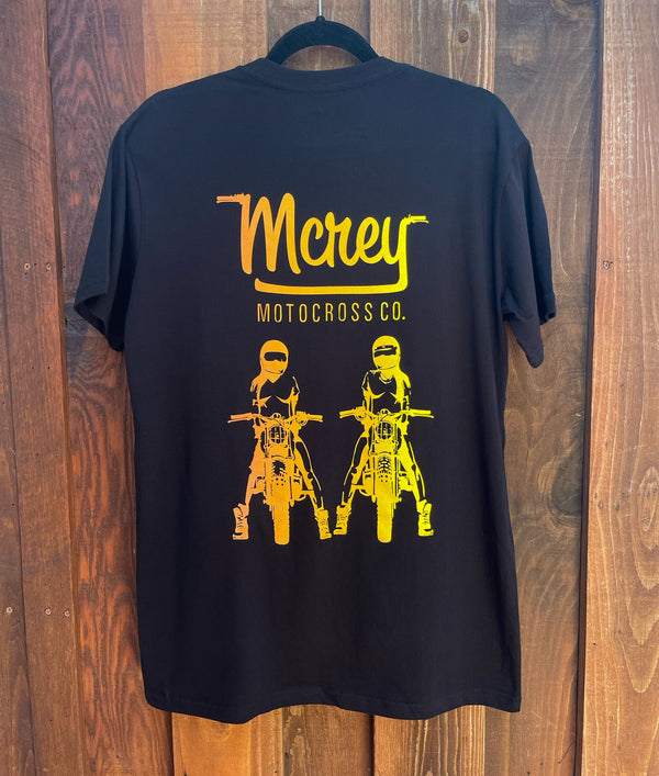Back of a unisex black shirt with two girls sitting on bikes in orange and yellow.