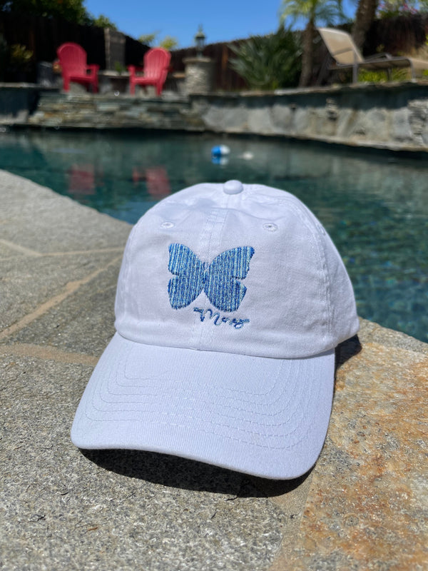 Youth white baseball-style cap with a blue-blend butterfly stitched on the front sitting next to a pool.