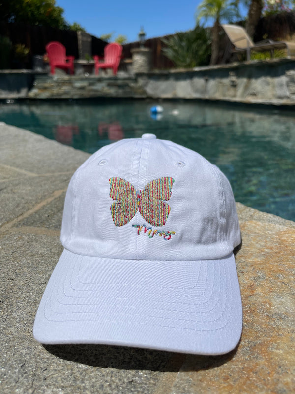Youth white baseball-style cap with a rainbow butterfly stitched on the front sitting next to a pool.